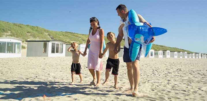 Eine Tour zum Strand ist für die ganze Familie fantastisch Mutter und Vater gehen Hand in Hand mit deren beiden Jungs am Strand. Vater hat ein großes Badetier unter dem Arm