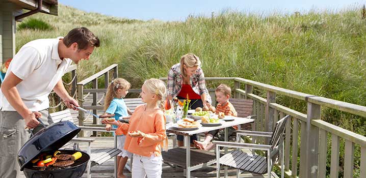 Die Terrasse Ihres Ferienhauses ist perfekt für gemütliche Familienessen und gute Grillabende Familie auf der Terrasse bei einem Ferienhaus. Der Vater händigt seiner Tochter etwas vom Grill, während der Rest der Familie im Hintergrund am Tisch sitzt