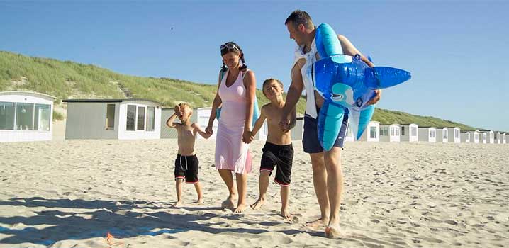 Mum and dad walking holding hands with their two boys on the beach. Dad is carrying a big water toy under his arm